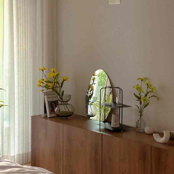 木目調のキャビネットと白いカーテンが調和するプランテリア空間、黄色い花と個性的な鏡が配置された自然な雰囲気のインテリア