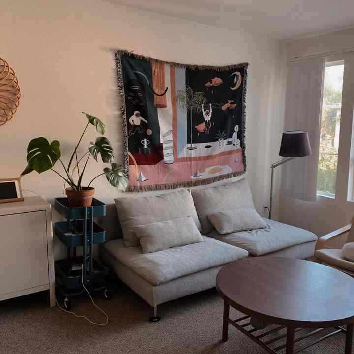 Natural-toned living room featuring a light gray 3-seat sofa, wooden round coffee table, a throw hung on the wall, and a Monstera plant creating a cozy atmosphere