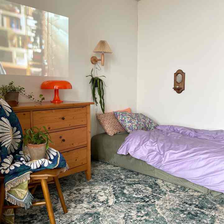 White-walled studio bedroom featuring wood-tone furniture, purple bedding, and floral pillows with a cozy atmosphere