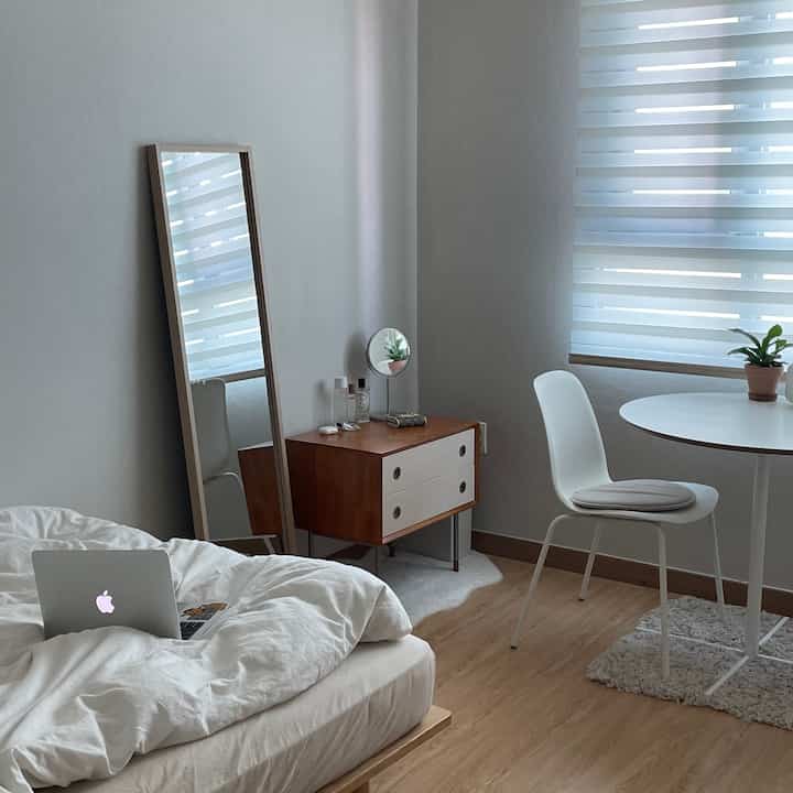 A wood-tone and white studio apartment featuring a bed, nightstand, and round table with a comfortable and clean atmosphere