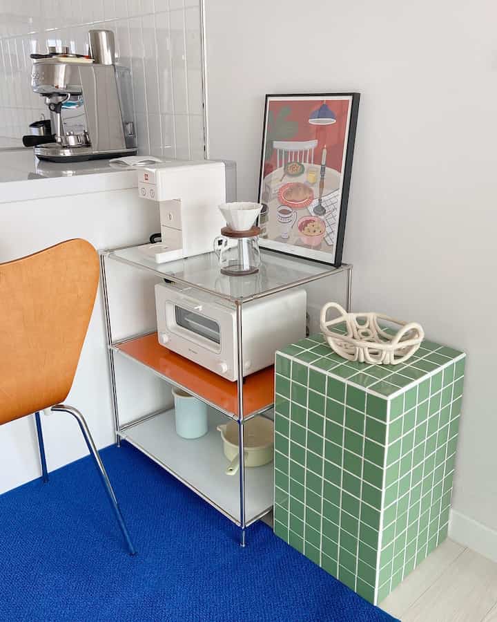 ブルーのラグと白い壁が調和するキッチン横のモダンなホームカフェ空間、サイドテーブルとコーヒーマシンが配置されている