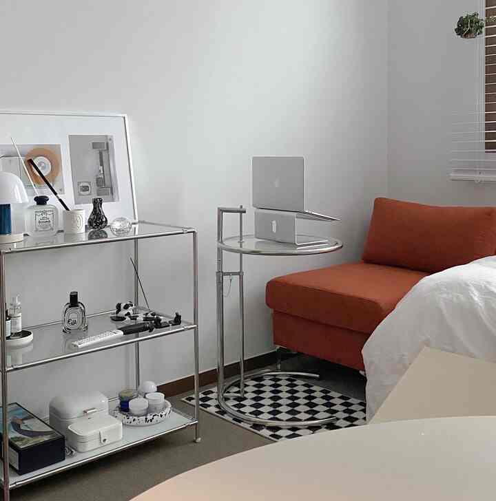 Living room with white walls and an orange sofa chaise, featuring stainless steel shelving and a modern round table in a clean interior