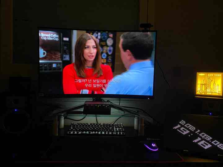 Dark-lit home office space with black desk featuring glass monitor stand, keyboard and mouse for remote work