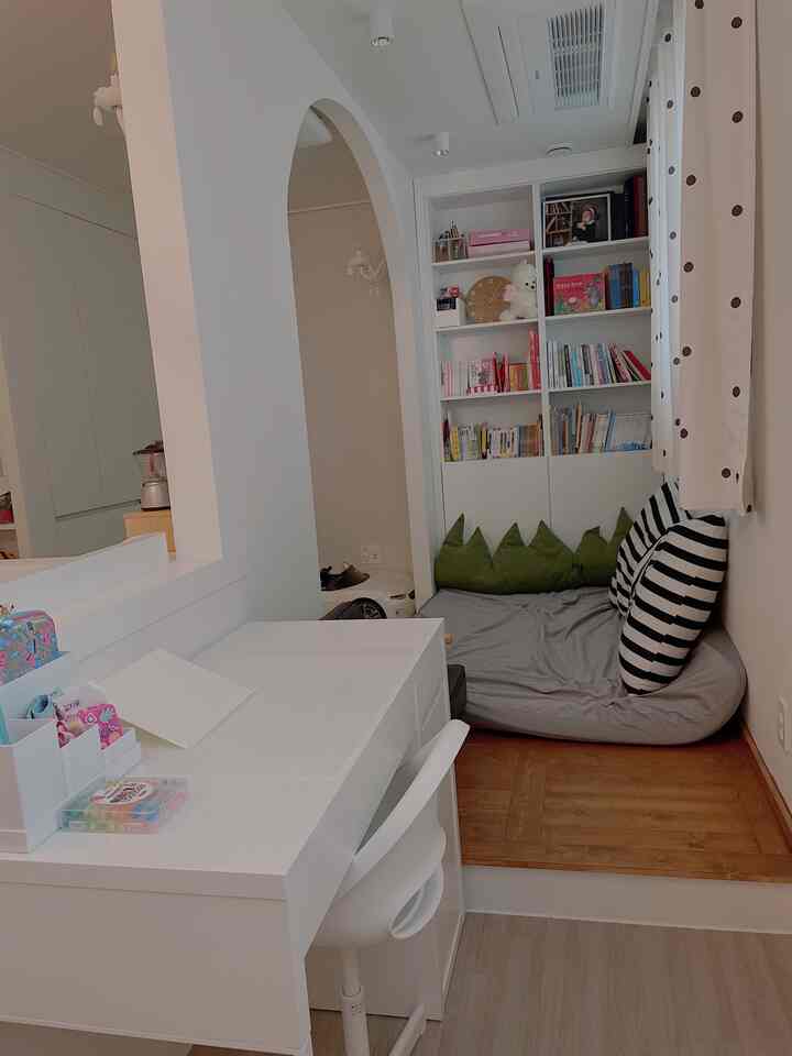 A compact kids' room reading nook with white and wood tones, featuring an arched room divider, desk, bookshelf, and cushions creating a cozy atmosphere