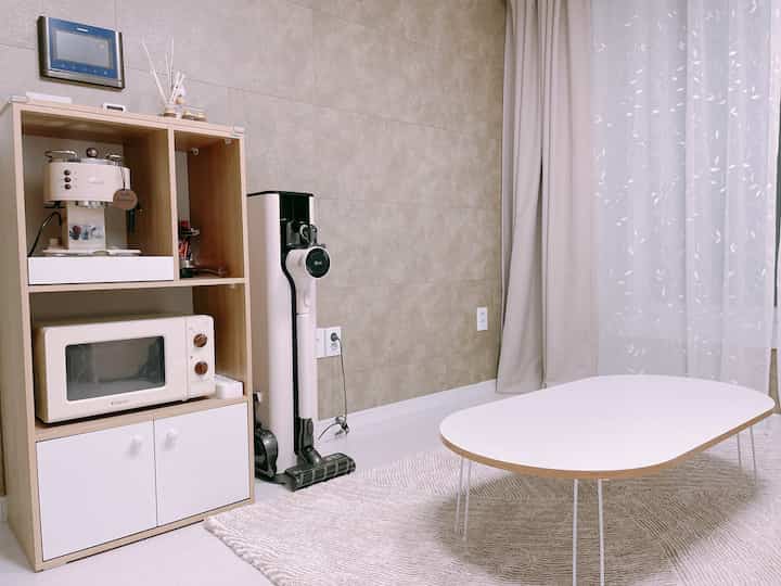 Beige-toned living room featuring a tidy kitchen shelf with semi-automatic coffee machine, vacuum cleaner, and round table arranged simply
