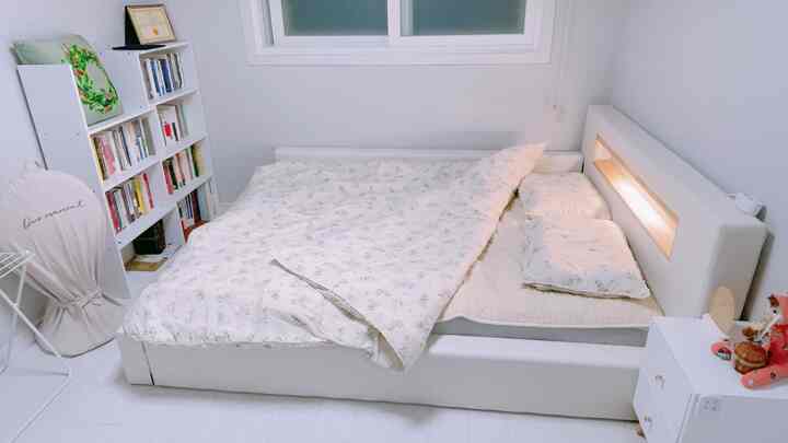 Calm white-toned bedroom featuring a white bed with floral duvet cover in center and a bookshelf on the left, cozy atmosphere