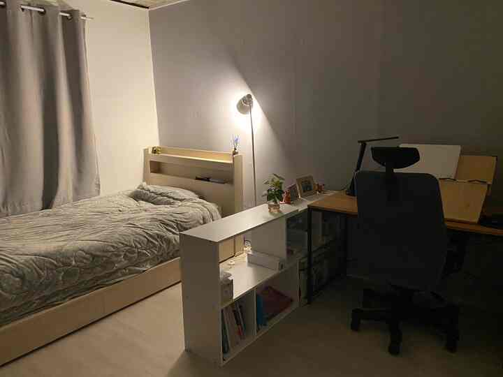 白い壁とベージュのベッド、デスクが置かれた一人暮らしの寝室兼ホームオフィスの空間