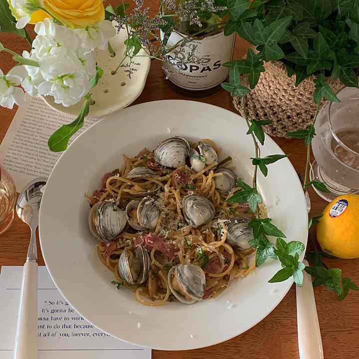 Natural wood table kitchen space featuring a white plate of clam pasta, with fragrant flowers and plants creating a cozy atmosphere