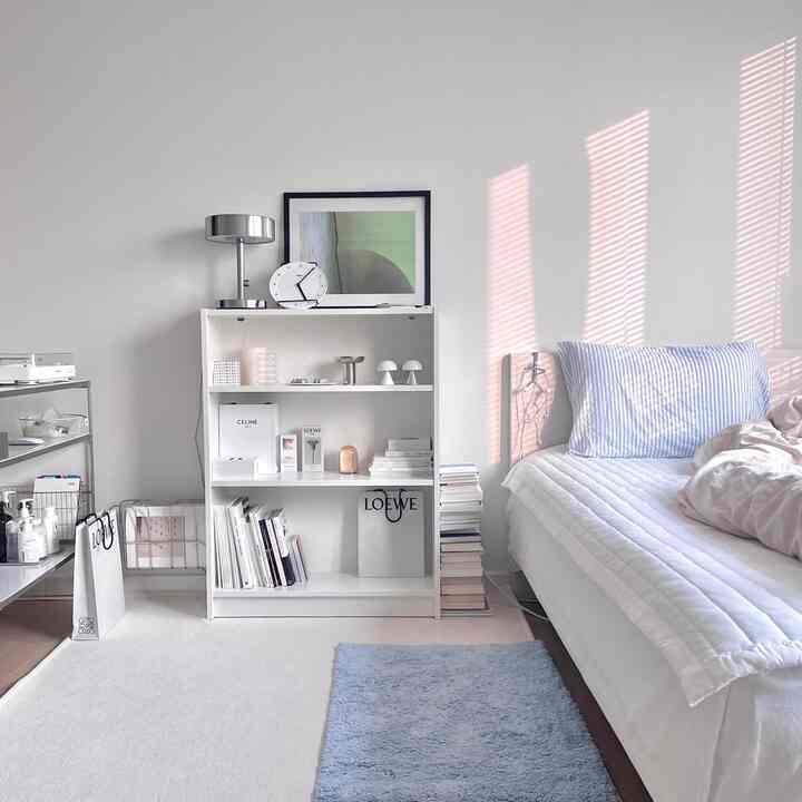Bright natural light fills a white-toned bedroom featuring a white shelf and blue rug in a simple, minimal setting