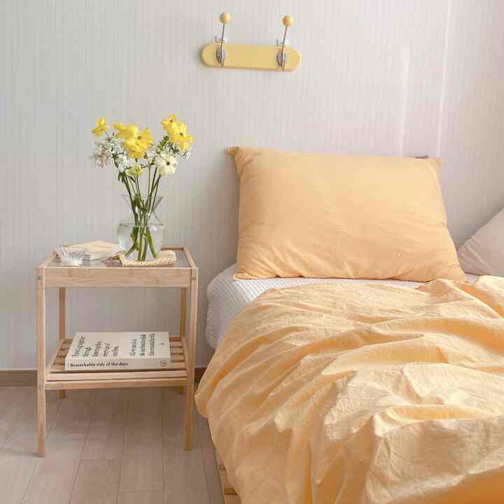 黄色と木目調の寝室、黄色い掛け布団カバーと枕、木製のナイトテーブルに花瓶が置かれた雰囲気のある空間