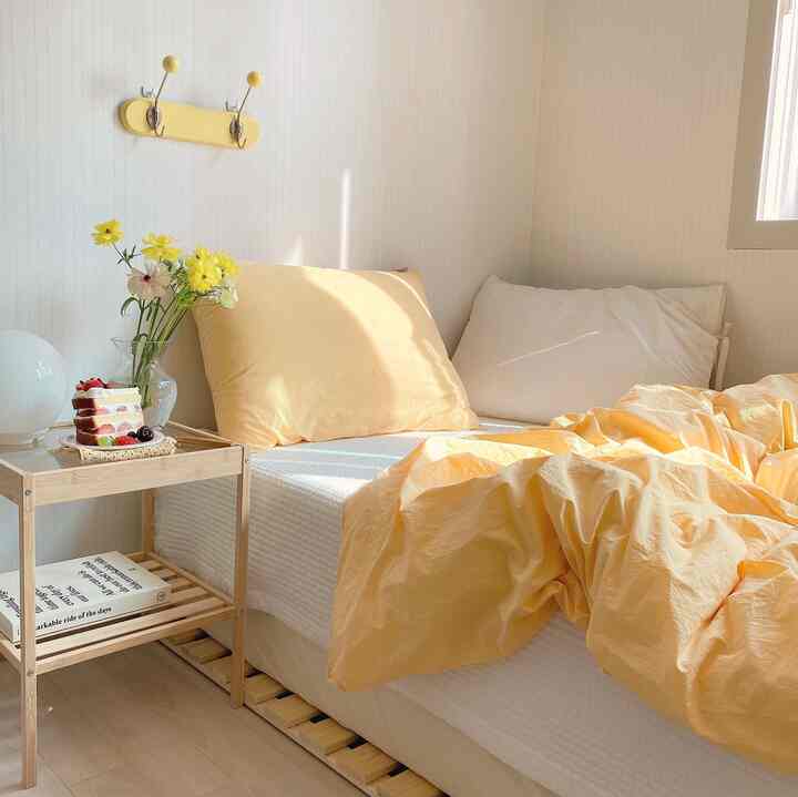 Cozy bedroom in warm yellow and white tones featuring bed and natural wood nightstand bathed in sunlight