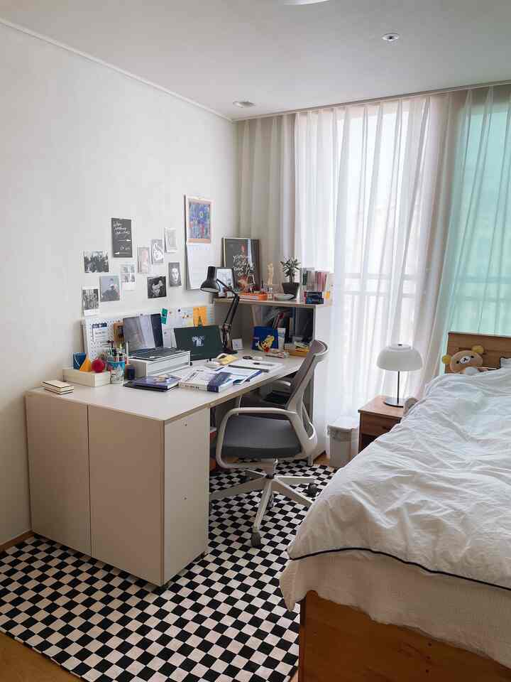 Bright natural-toned bedroom and home office featuring a large desk, ergonomic chair, bed, and checkered rug, with a clean cozy atmosphere
