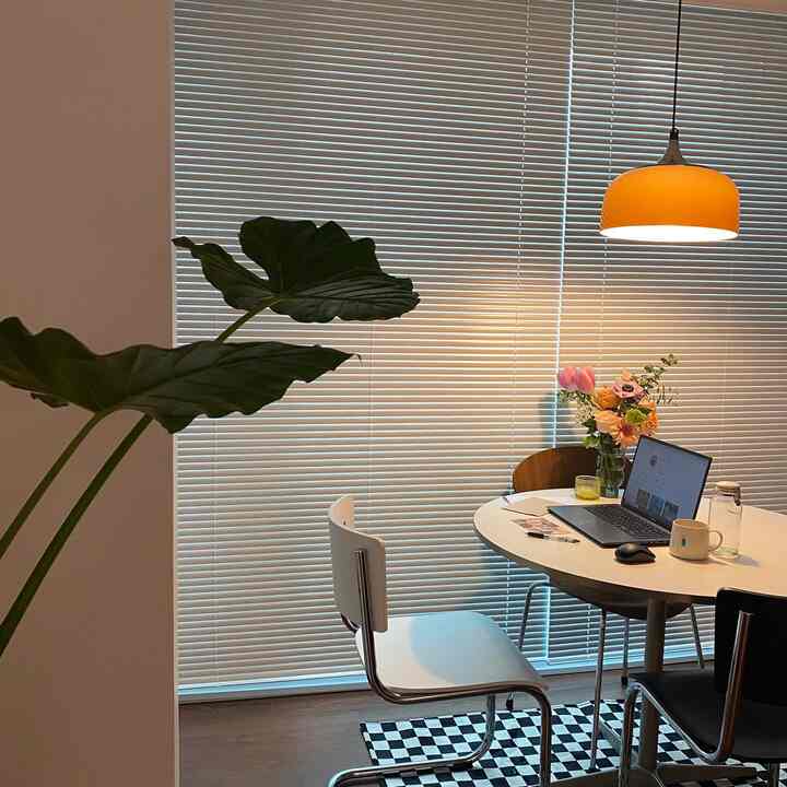 Natural-toned home office with yellow pendant light and white round dining table by window blinds