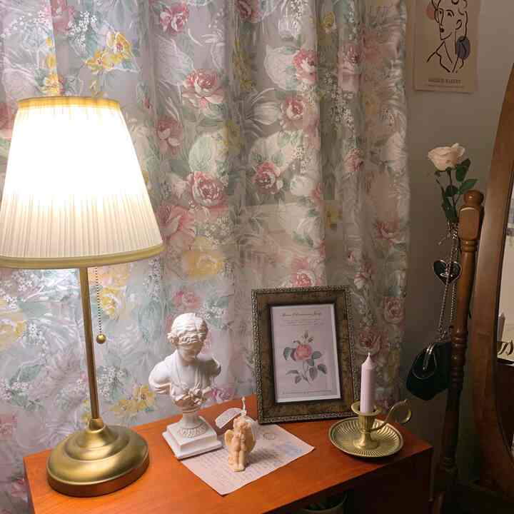 ゴールドとブラウンのトーンが特徴の小さなテーブルに卓上ランプ、石膏像、ヴィンテージキャンドルホルダーが配置された暖かみのある寝室の空間