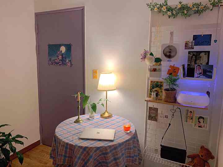 A home cafe space with white walls and brown wood flooring featuring a round table, vintage-style lamp, plants, and a metal partition creating a cozy atmosphere