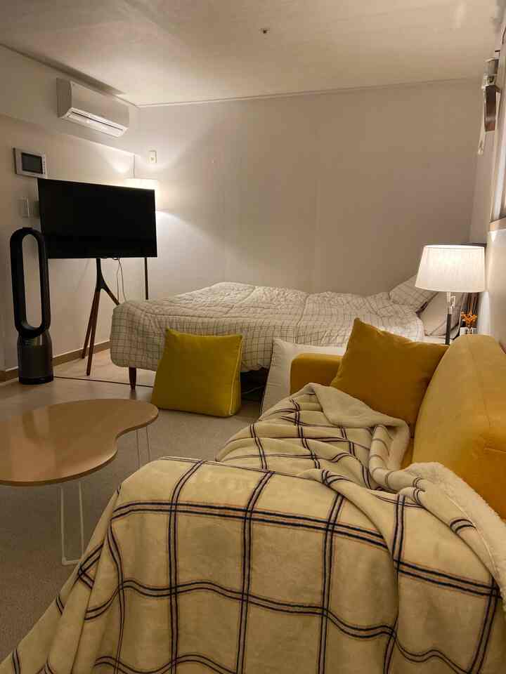 Yellow and ivory toned living and bedroom space featuring a yellow sofa, white checkered bed, cozy lighting, and wood tone furniture