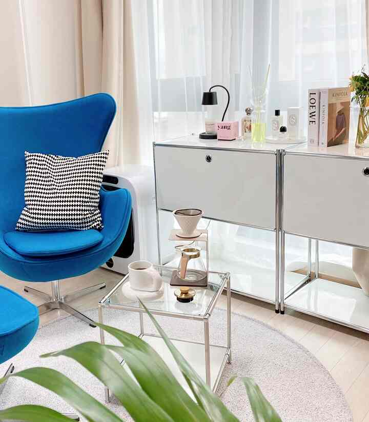 A white and blue toned studio living room featuring an Egg Chair, modular furniture, and a transparent glass coffee table with a modern vibe