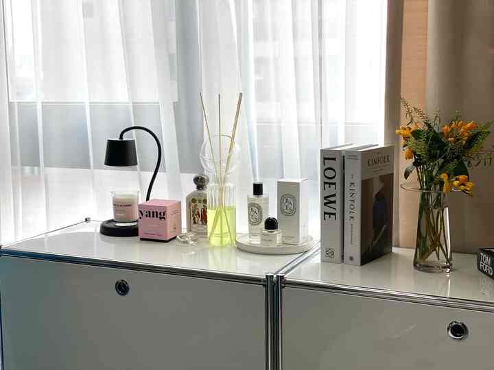 Studio apartment interior with white curtains and white storage cabinet, featuring vases, candles, and diffuser on a neat surface
