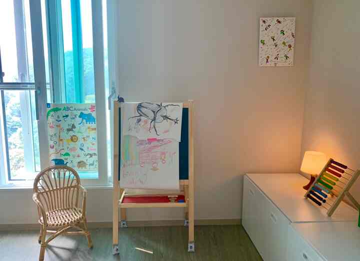ナチュラルな木目調と白が調和した子供部屋、ラタンチェアと子供の絵が掛かったイーゼル、照明と収納ベンチがある雰囲気のあるインテリア