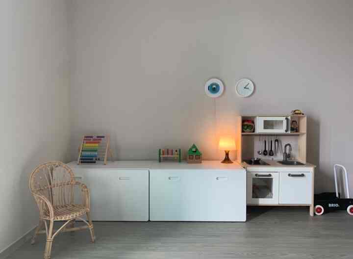 白とナチュラルトーンの子供部屋、玩具収納とキッチンおもちゃセット、テーブルランプが配置された印象的な空間