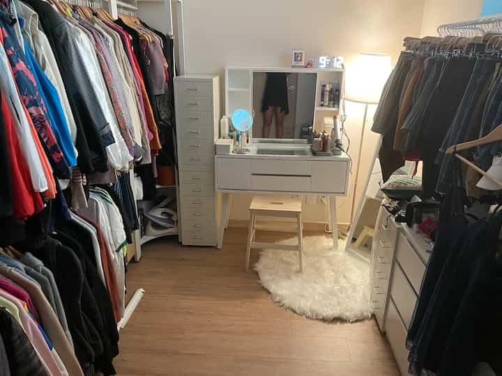 A cozy walk-in closet in white and wood tones featuring clothing racks, a vanity table, rug, and floor lamp