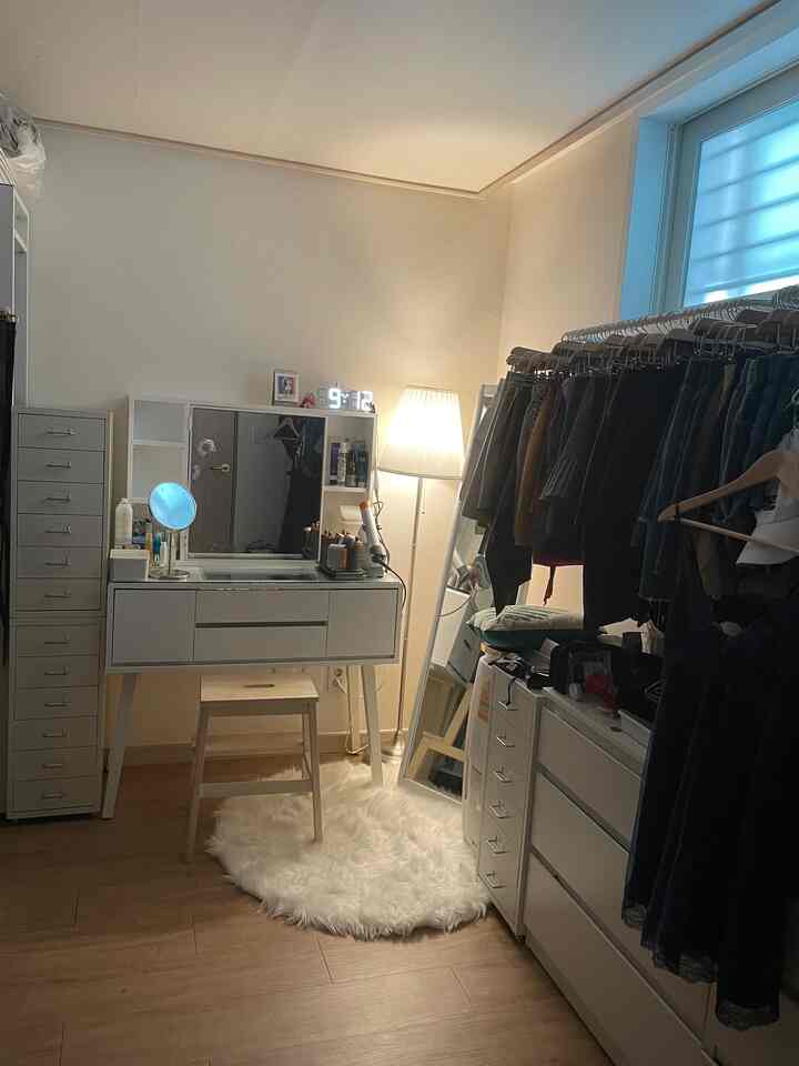 A cozy studio apartment with white and wood tones featuring a vanity, dresser, floor lamp and clothing rack in a compact space