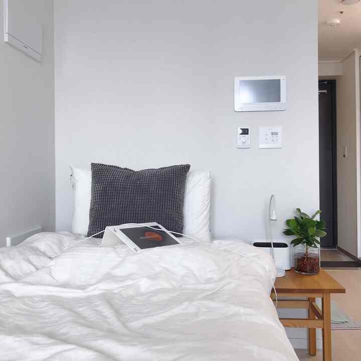 Minimal studio bedroom with white bedding and wood tone bedside table featuring a tablet stand and green plant