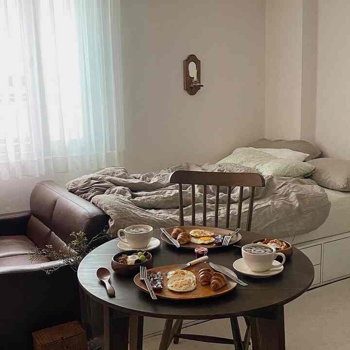 ナチュラルなベージュ調のワンルーム寝室、木製家具とヴィンテージレザーソファ、ホームカフェのブランチテーブルが配置された温かみのある空間