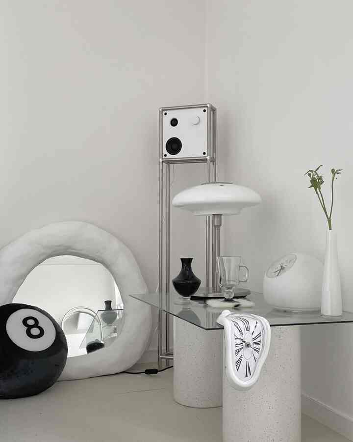 アイボリーとホワイトトーンのモダンな空間、ユニークなガラステーブルと幾何学的なランプ、アート風の時計や鏡が配置された洗練されたインテリア