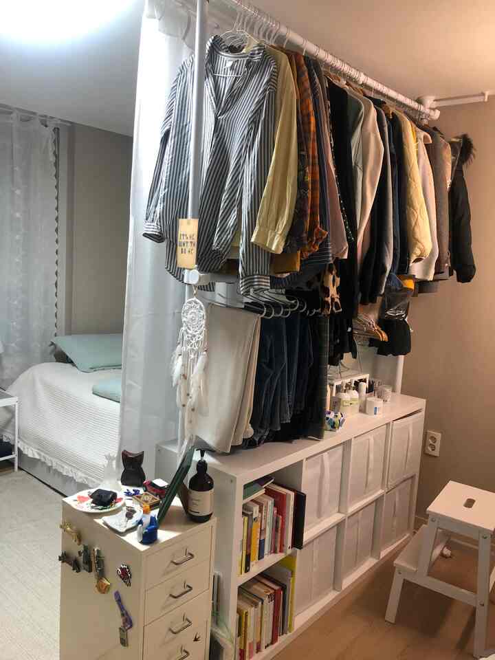 ホワイトとナチュラルカラーのコンパクトな寝室、中央にドレスルーム機能の衣類ラックと収納家具が配置された雰囲気のある空間