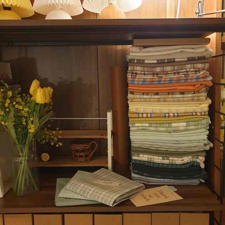 ブラウンの木製棚にカラフルなファブリックの枕カバーがきれいに積まれ、左側には黄色いチューリップが入ったガラスの花瓶が置かれた整理された空間