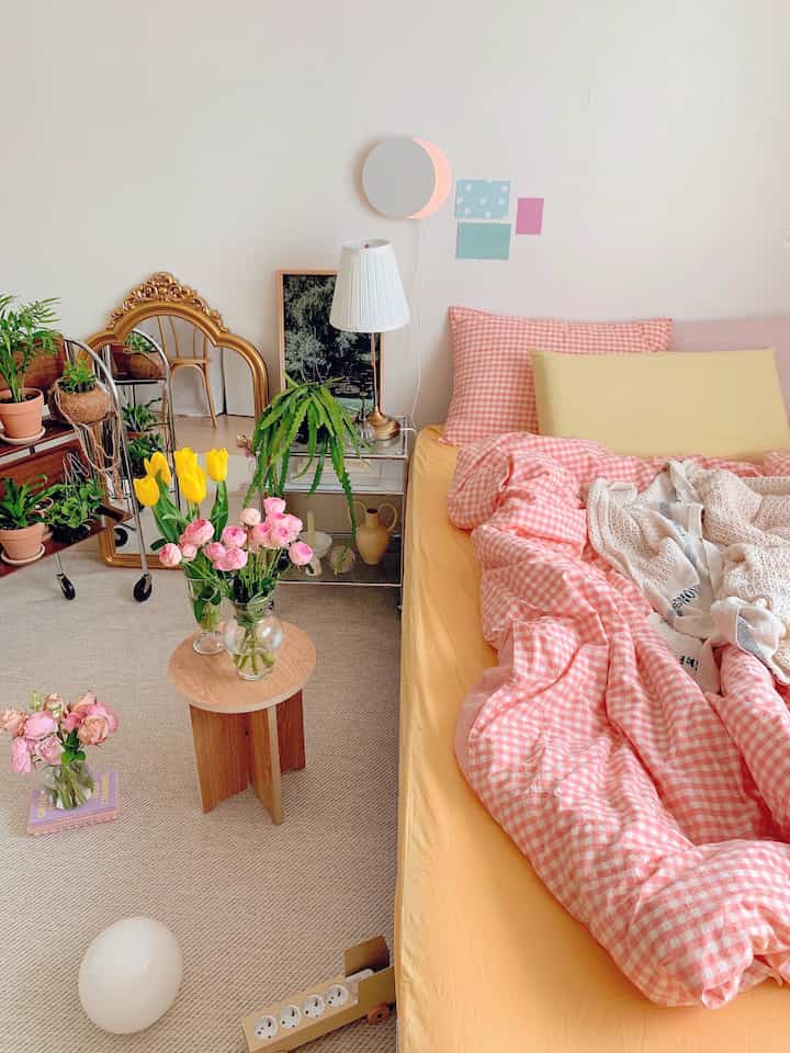 Bedroom with white walls and wood tone furniture, featuring pink gingham bedding and vibrant plants in a cozy setting