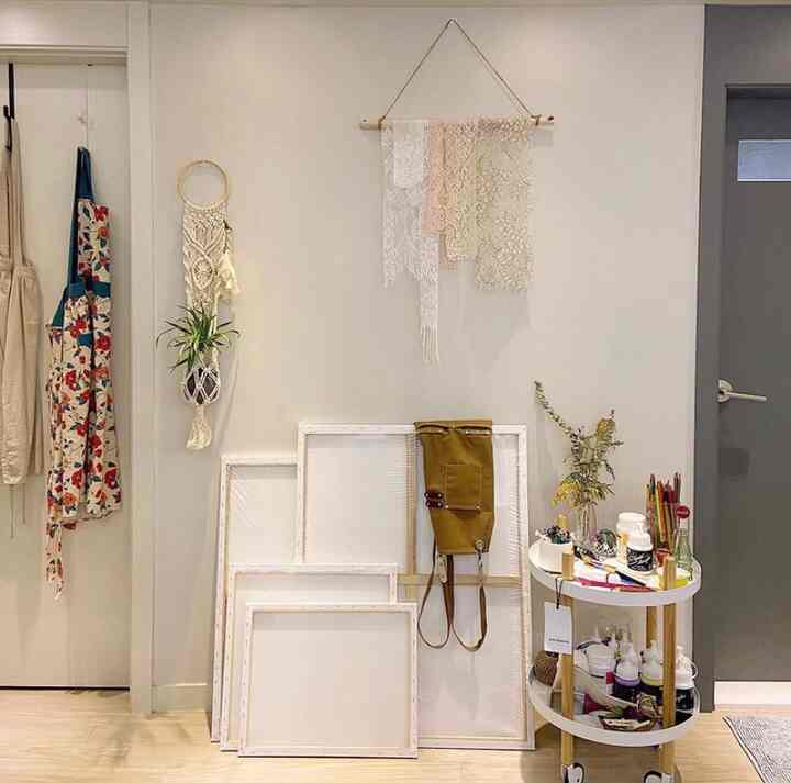 Natural tone small workspace featuring lace wall hanging, unframed canvases, aprons, and a kitchen cart neatly arranged