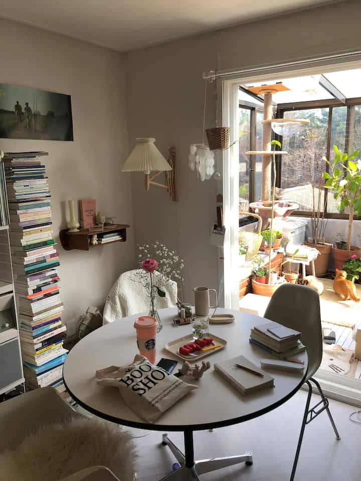 Ivory and white toned workspace adjacent to veranda, featuring round table, bookshelf, numerous plants and cats in a calm cozy atmosphere