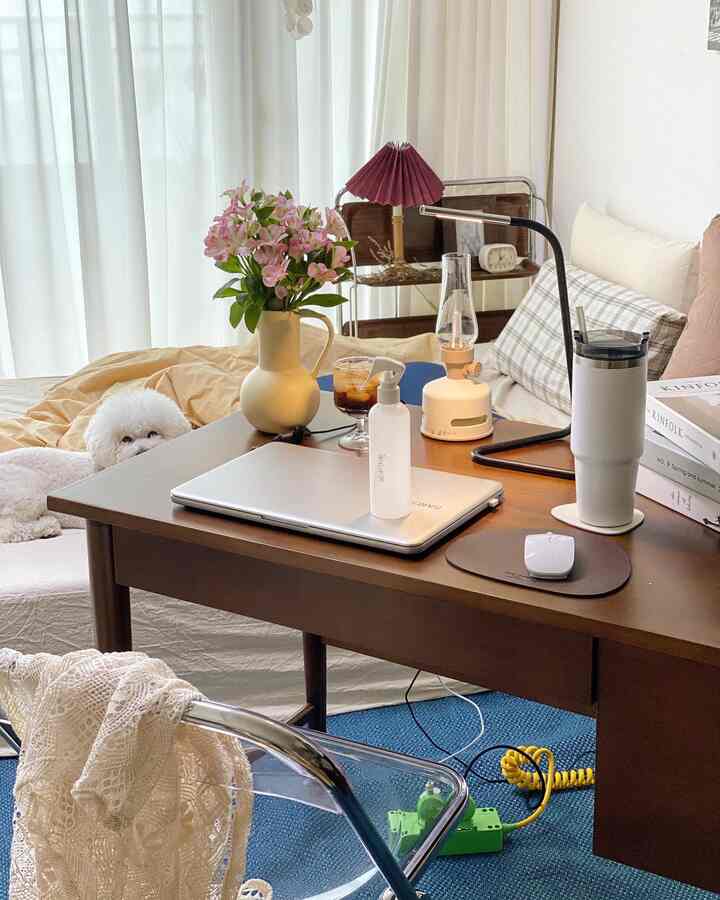 ベージュと木目調の寝室ホームオフィス。デスクの上にノートパソコンと花瓶があり、小さなペットがいる雰囲気のある空間