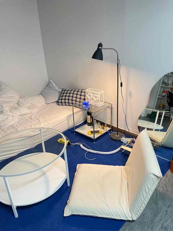 Living and bedroom space with white walls and bedding, blue rug flooring, simple interior featuring a module nightstand and floor lamp