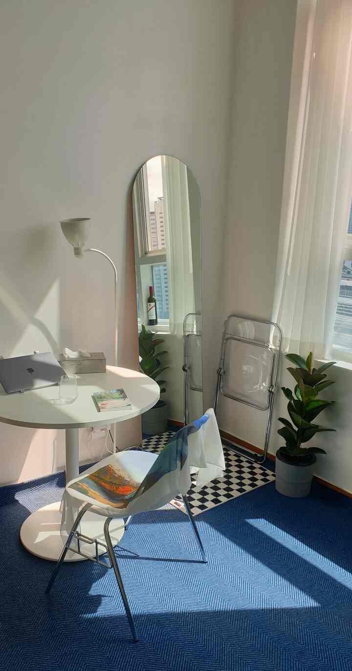 白い壁とネイビーのラグが敷かれたワンルーム、丸いテーブルと椅子、全身鏡とフロアランプが配置されたモダンで落ち着いた雰囲気の空間