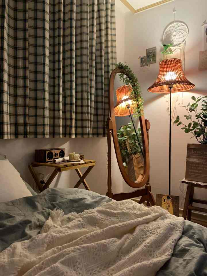 ナチュラルな木目調とグリーンの植物が調和した小さな寝室、木製の全身鏡とフロアランプが温かい雰囲気を演出