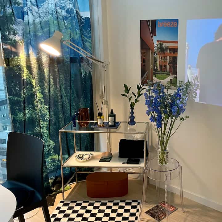 A natural living room with blue mountain forest curtains as background, featuring a transparent stool, modular shelf, blue flowers, and wall lamp creating a cozy atmosphere