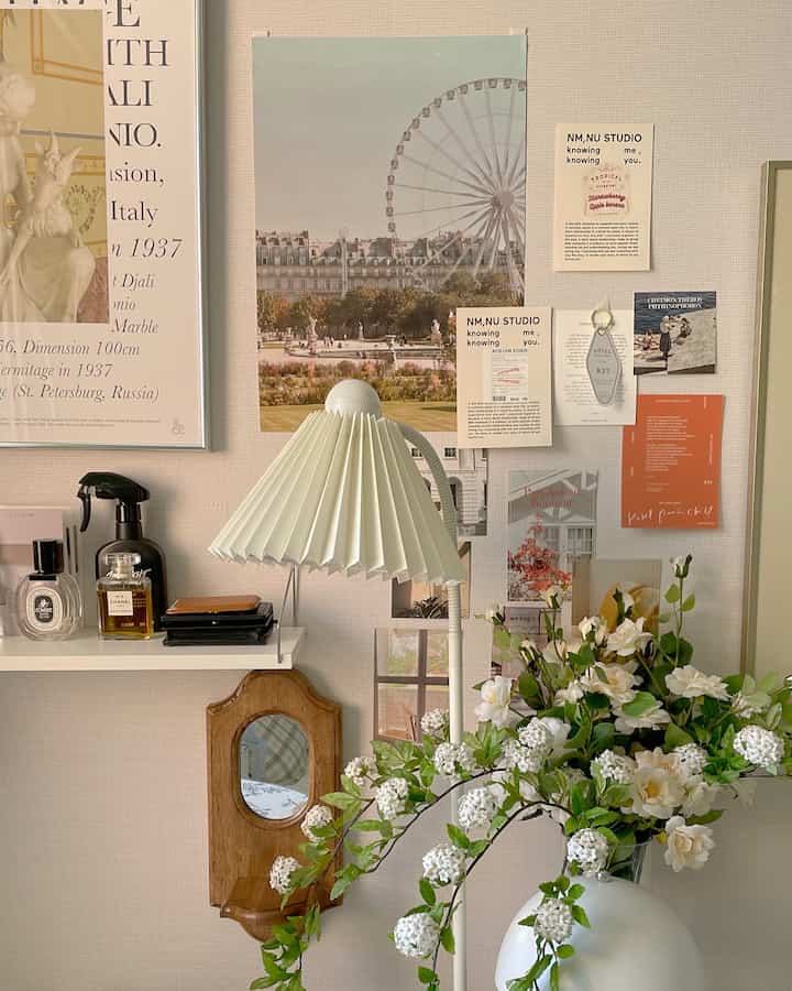 White and natural toned studio workspace featuring wall shelf, multiple posters, artificial flowers, and a floor lamp creating cozy atmosphere
