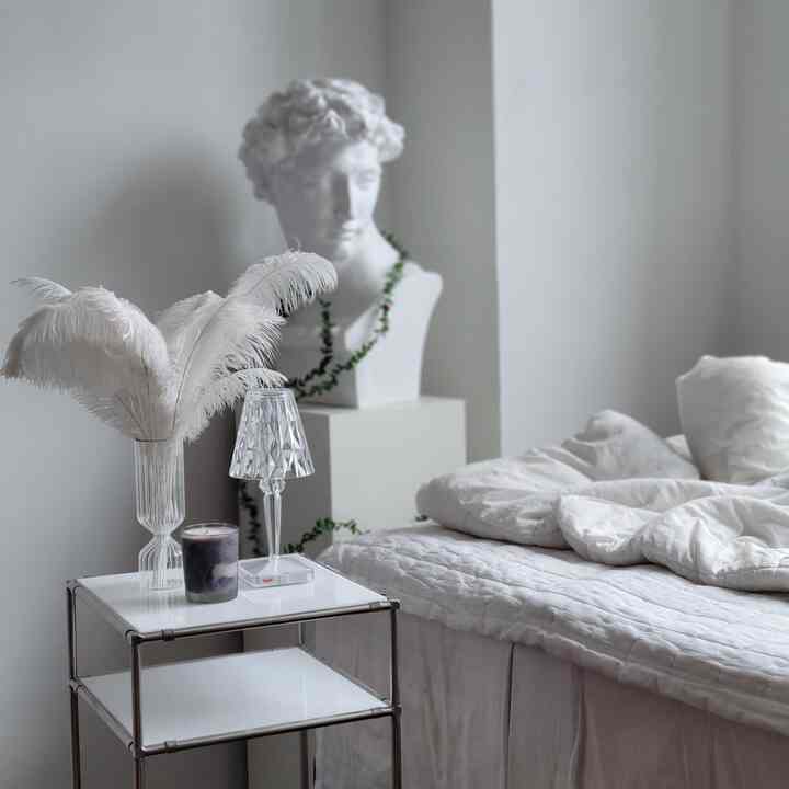 White-toned bedroom featuring a nightstand with feather decor and a plaster bust, with soft bedding creating a modern calm space