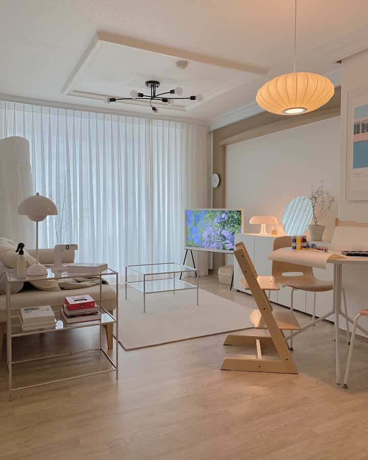 Modern living room with white curtains and natural wood tone furniture, featuring transparent coffee table and rug creating a stylish atmosphere