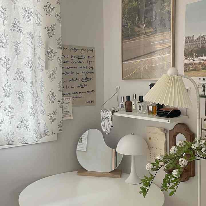ホワイトとベージュの色調が特徴のワンルームワークスペースで、丸テーブルと花柄のカーテン、鏡や多様な照明が配置された雰囲気のあるインテリア