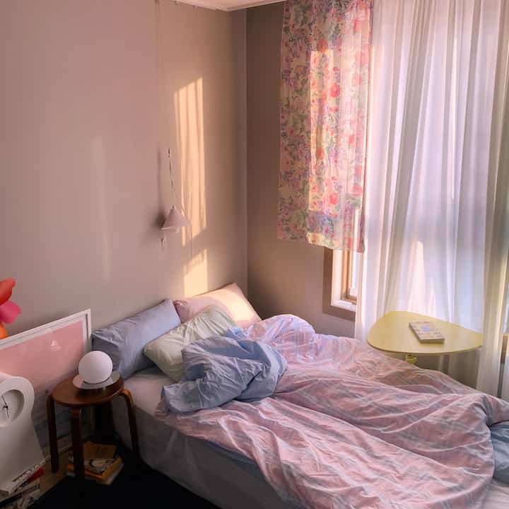 パステルカラーと白色が調和したワンルーム寝室、窓辺の花柄カーテンとベッドの掛け布団カバーが温かみのある雰囲気を演出している