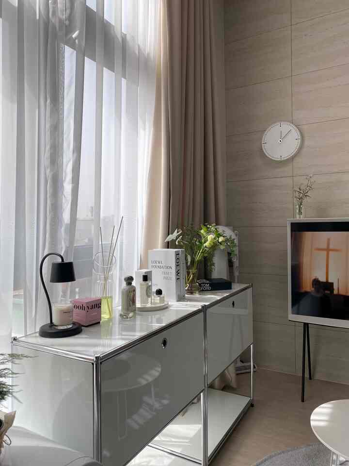 明るいベージュのカーテンと白いチェストが配置された白を基調としたワンルームのリビング、ミニマルな壁時計とテーブルランプがアクセントのインテリア