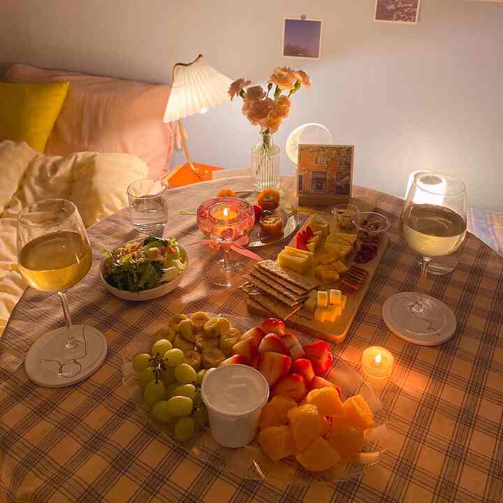 A soft pink and light brown toned studio apartment dining space featuring a checkered table with fruits, candles, and a vase creating a cozy home cafe atmosphere