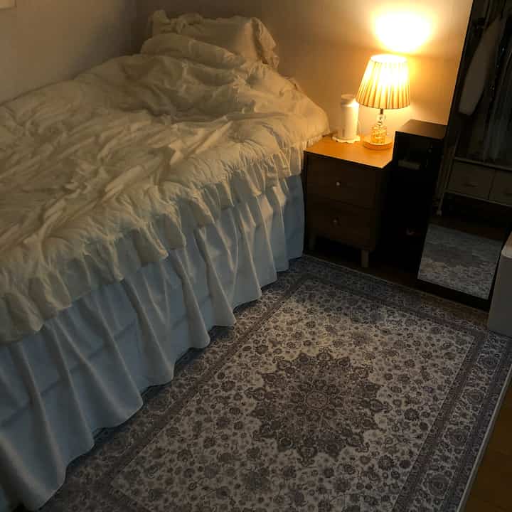 温かみのある照明と白い寝具がポイントの小さなワンルーム寝室、木目調のナイトテーブルと柄のラグが調和した雰囲気のあるインテリア