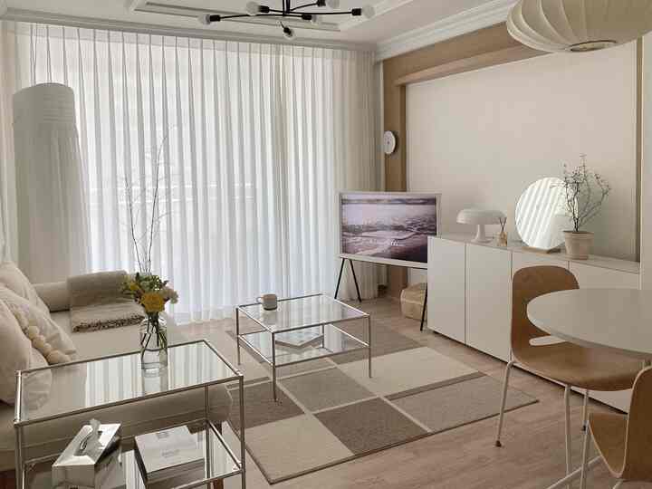 White and brown toned living room featuring beige sofa, glass tables, long chiffon curtains, and a neat rug creating a warm atmosphere