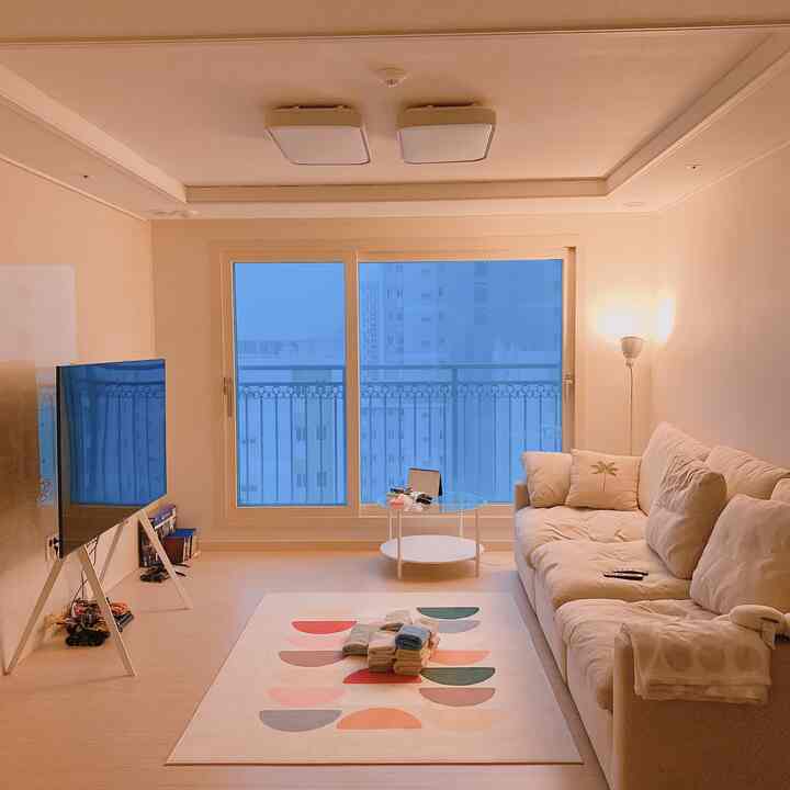 暖かいベージュトーンのリビング、大きなベージュのソファと丸いコーヒーテーブル、テレビ台とフロアランプが配置された落ち着いた空間