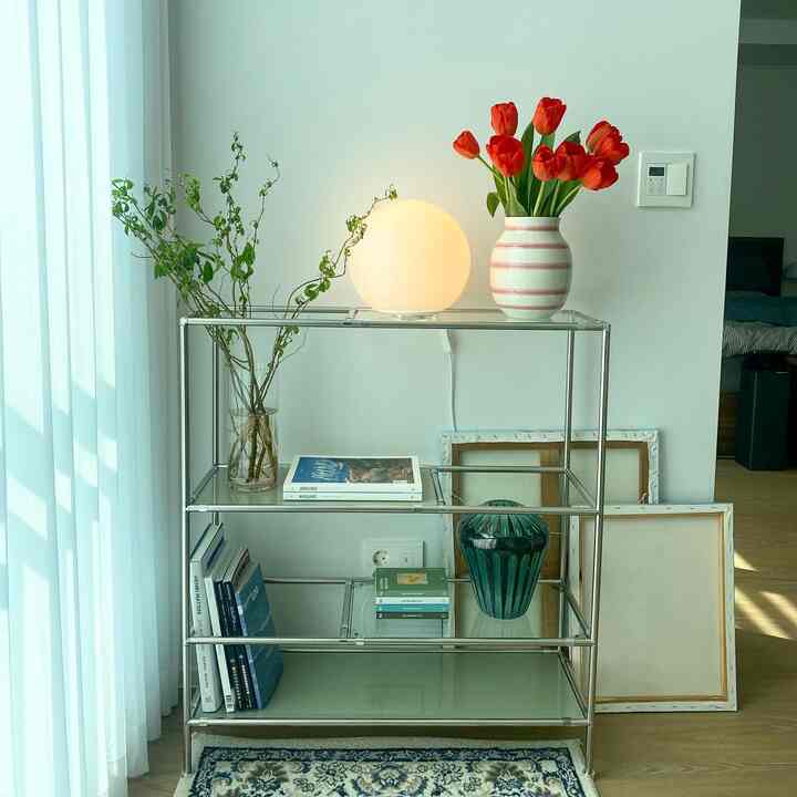 Natural toned space with white walls and curtains, featuring a modular glass shelf with a lamp, red tulip vase, and plants, creating a simple study area